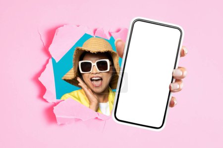 Foto de Oferta móvil. Mujer asiática emocionada en gafas de sol y sombrero de verano que muestra el teléfono inteligente, pantalla de maqueta vacía a través del agujero rasgado en el fondo de papel rosa, dispositivo de presentación o nueva aplicación. Collage. - Imagen libre de derechos