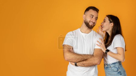Téléchargez les photos : Un homme européen barbu et heureux millénaire croise les bras et sourit comme une femme murmure dans son oreille, à la fois vêtu de chemises blanches et de jeans sur un fond orange uni, panorama - en image libre de droit