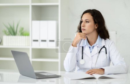 Téléchargez les photos : Pensive belle femme millénaire portant manteau médical médecin généraliste assis au bureau en face de l'ordinateur portable dans son bureau à la clinique, regardant l'espace de copie. Assurance maladie - en image libre de droit