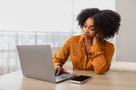 Téléchargez les photos : Une jeune femme afro-américaine triste et réfléchie aux cheveux bouclés, portant une chemise en moutarde, s'appuie sur sa main tout en utilisant un ordinateur portable, peut-être en travaillant ou en étudiant au bureau à domicile - en image libre de droit