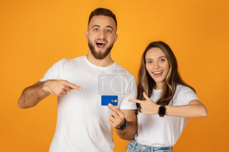 Photo for Enthusiastic european young couple pointing at a credit card held by the woman, both wearing white t-shirts and smiling, against a monochromatic orange background, studio - Royalty Free Image