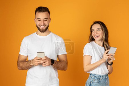 Photo for Happy european young man and woman look at smartphone, chatting in social networks, enjoy surfing, isolated on orange background studio. App and fun, gadget for communication - Royalty Free Image