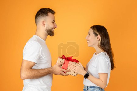 Téléchargez les photos : Une femme européenne souriante remet un cadeau festif avec un ruban rouge à un homme heureux, portant tous deux des t-shirts blancs et se regardant, sur un fond orange - en image libre de droit