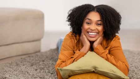 Foto de Radiante feliz millennial mujer afroamericana con el pelo rizado natural acostado en el suelo, descansando su barbilla sobre una almohada, sonríe brillantemente a la cámara en una pose relajada, panorama - Imagen libre de derechos