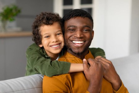 Photo for Happy black boy with curly hair joyfully hugs his father from behind, both sharing radiant smiles in comfortable home setting, man sitting on couch - Royalty Free Image
