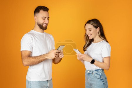 Foto de Comprometidos en la comunicación moderna, los jóvenes europeos sonríen mientras usan sus teléfonos inteligentes, posiblemente enviando mensajes de texto o navegando por las redes sociales, sobre un fondo naranja. - Imagen libre de derechos
