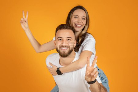 Foto de Una alegre pareja joven europea posa juguetonamente con el hombre cargando a la mujer en su espalda, ambos mostrando signos de paz, sobre un fondo naranja. Diversión, juego y tiempo libre - Imagen libre de derechos
