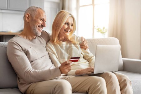 Photo for Happy loving caucasian retired man and woman banking online together, using laptop computer pc and red plastic bank credit card, sitting on couch at home, copy space. Retail, ecommerce, virtual bank - Royalty Free Image