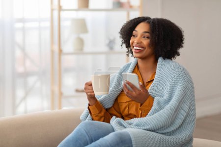 Photo for A joyful woman with natural curly hair, wrapped in a knitted blue blanket, laughs while looking at her smartphone in one hand and holding a mug in the other, seated on a couch - Royalty Free Image