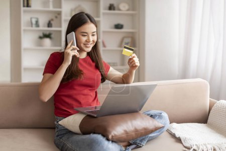 Photo for Smiling young asian woman holding credit card and talking on cellphone while making online purchase on her laptop, happy korean lady comfortably sitting on couch in living room at home - Royalty Free Image