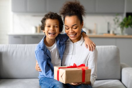 Téléchargez les photos : Mère noire rayonnante et son fils préadolescent ravi partagent un moment de bonheur avec une boîte cadeau, enveloppée d'un ruban rouge, assis sur le canapé - en image libre de droit