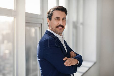 Téléchargez les photos : Homme d'affaires caucasien souriant avec une moustache, vêtu d'un blazer bleu et d'une chemise blanche, se tient avec confiance avec les bras croisés dans un intérieur de bureau moderne avec de grandes fenêtres - en image libre de droit