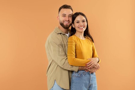 Photo for Happy young caucasian man hugging his girlfriend posing on orange background in studio, smiling to camera. Millennial family couple embracing for photo. Love and romantic date, Valentines day - Royalty Free Image