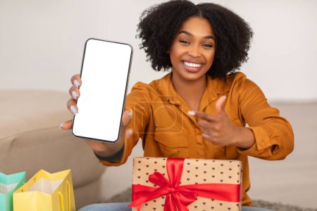 Foto de Emocionada mujer afroamericana milenaria sonriente mostrando una pantalla de teléfono inteligente en blanco y señalándola con una sonrisa, sentada en el suelo cerca de una caja de regalo y bolsas de colores - Imagen libre de derechos