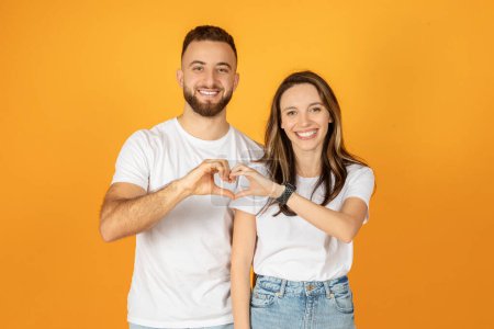 Photo for A smiling millennial european man and woman in white shirts make a heart shape with their hands together, exuding affection and happiness against a warm orange background - Royalty Free Image