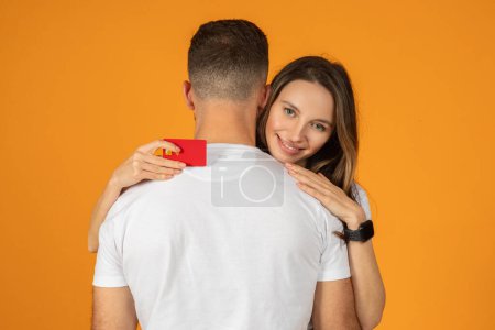 Téléchargez les photos : Une femme européenne souriante tenant secrètement une carte de crédit rouge derrière un dos d'homme crée une scène de surprise ou de cadeau, les deux portant des hauts blancs décontractés avec un fond orange - en image libre de droit