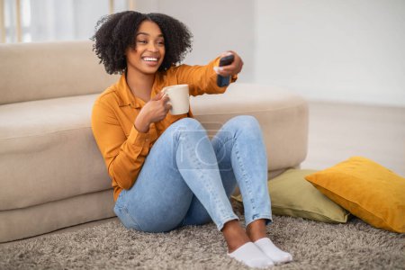 Téléchargez les photos : Radieuse femme afro-américaine heureuse assise sur le sol avec une tasse à la main, souriante lorsqu'elle utilise une télécommande, profitant probablement d'un moment de détente à la maison seule, pleine longueur - en image libre de droit