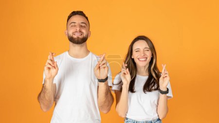Téléchargez les photos : Joyeux couple européen avec les yeux fermés, doigts croisés pour la bonne chance, souriant et espérant, portant des t-shirts blancs décontractés, debout sur un fond orange vif - en image libre de droit