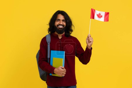 Foto de Alegre estudiante hindú con mochila y un montón de libros que muestran la bandera de Canadá sobre fondo de estudio amarillo, espacio para copiar. Joven positivo oriental estudiando en el extranjero. Educación en el extranjero - Imagen libre de derechos