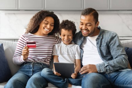 Foto de Alegre familia negra de compras en línea con la tableta digital y la tarjeta de crédito en el hogar, padres afroamericanos felices y su hijo pequeño que se relaja en el sofá con el gadget moderno, comprando en Internet - Imagen libre de derechos