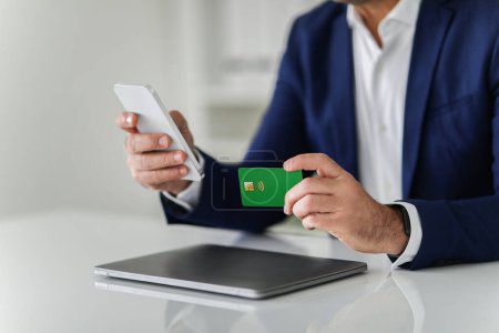 Photo for Close-up of caucasian man in a business suit performing a contactless payment with a green credit card and smartphone over a closed laptop in an office. Work, business gadget, app - Royalty Free Image