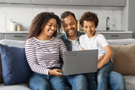 Photo for Cheerful black family having fun with laptop at home, watching photos together. African american parents and their little son using computer while sitting on couch in living room, free space - Royalty Free Image