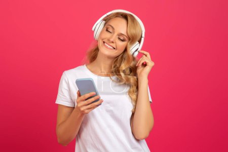 Foto de Ocio musical. Mujer rubia joven feliz con teléfono inteligente y auriculares inalámbricos relajante, escuchando música en aplicación móvil sobre fondo rosa en el estudio. Gadgets, tecnología - Imagen libre de derechos