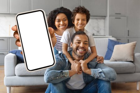Foto de Alegre familia afroamericana de tres personas mostrando un teléfono inteligente en blanco con pantalla blanca para maqueta mientras se relaja en casa, padres negros felices e hijo pequeño recomendando una nueva aplicación móvil, maqueta - Imagen libre de derechos