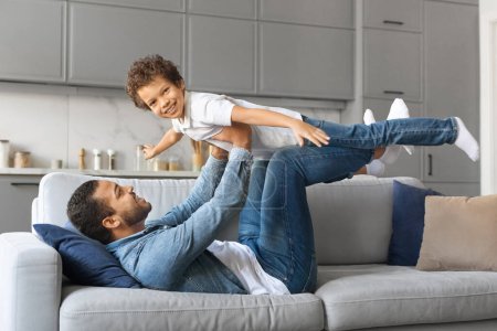 Photo for Family Fun. Happy black father playing with little son at home, lying on couch and carrying his male kid like airplane, holding boy on hands, child pretending flying spreading hands as wings - Royalty Free Image