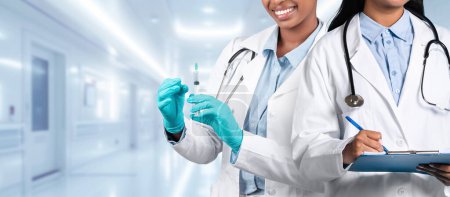 Foto de Una doctora con una bata blanca que prepara una vacuna con una jeringa está acompañada por un colega sonriente con un portapapeles, ambos listos para la atención del paciente en un pasillo del hospital. - Imagen libre de derechos