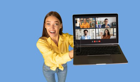 Foto de Una mujer milenaria europea alegre en una blusa amarilla extiende su brazo para presentar una pantalla del ordenador portátil que muestra una videollamada de grupo amistosa, fijada sobre un fondo azul brillante, panorama - Imagen libre de derechos