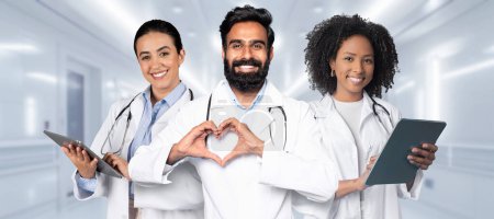 Photo for A diverse team of two women and one man healthcare professionals, one with a tablet and another making a heart shape with hands, in a hospital corridor. Professional cardio help - Royalty Free Image