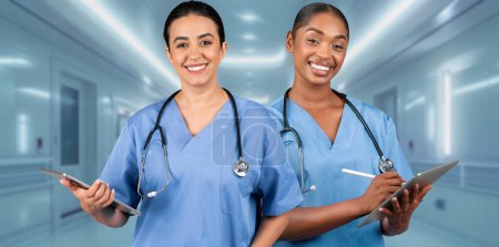 Téléchargez les photos : Deux infirmières afro-américaines souriantes en blouse bleue tenant des tablettes numériques se tiennent en confiance dans un couloir d'hôpital, représentant un environnement de santé moderne - en image libre de droit