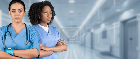 Zwei seriöse europäisch-afrikanisch-amerikanische Krankenschwestern in blauem Peeling mit verschränkten Armen und Stethoskopen stehen entschlossen auf einem Krankenhausflur und spiegeln Stärke und Professionalität wider.