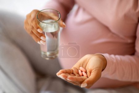Foto de Vitaminas y suplementos prenatales. Mujer embarazada negra irreconocible que toma pastillas y vaso de agua, inyección recortada de joven embarazada que usa medicamentos para un embarazo saludable, primer plano - Imagen libre de derechos