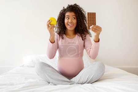 Téléchargez les photos : Joyeux noir femme enceinte assise sur le lit à la maison, comparant la pomme à la barre de chocolat, heureuse femme enceinte afro-américaine incarnant les choix de grossesse et les envies, espace libre - en image libre de droit