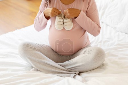 Photo for Pregnant lady sitting cross-legged on bed and holding pair of small white baby shoes over her belly, unrecognizable young african american female anticipating motherhood, cropped shot - Royalty Free Image