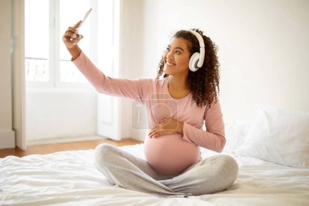 Photo for Radiant black pregnant woman with headphones enjoying music and taking selfie, happy african american expectant mother capturing cheerful moment on smartphone camera while sitting on bed at home - Royalty Free Image