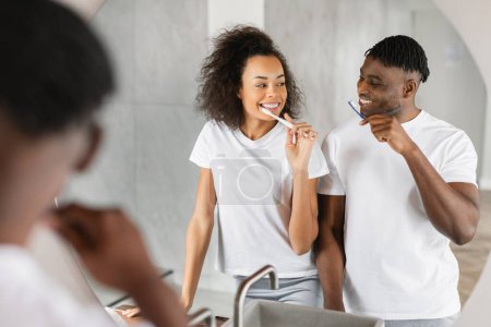 Foto de Cuidado dental. Loving Black Millennial Couple Sharing Smiles as They Brushing Teeth Near Mirror, Enjoying Happy and Health Conscious Morning at Home Bathroom (en inglés). Enfoque selectivo - Imagen libre de derechos