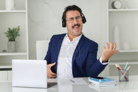 Photo for A lively caucasian businessman in a blue suit joyfully sings along to music, wearing headphones at his modern office desk, portraying a break or stress relief moment during work - Royalty Free Image