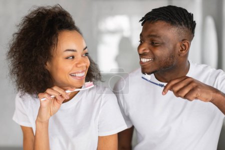 Téléchargez les photos : Gros plan de joyeux couple afro-américain se brossant les dents ensemble en profitant des rituels dentaires matinaux dans la salle de bain à la maison. Petit ami noir et petite amie s'occupant de l'hygiène buccodentaire tenant des brosses à dents - en image libre de droit
