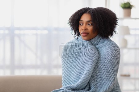 Photo for Pensive focused African American woman with curly hair wrapped snugly in a blue knitted blanket, looking off into the distance in a bright, serene room, at free space at home - Royalty Free Image