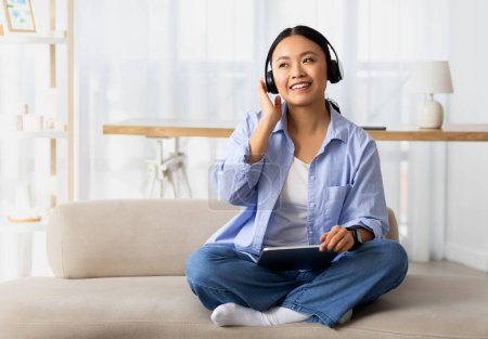Foto de Mujer coreana bastante joven feliz usando auriculares inalámbricos y tableta digital, sentado en el sofá en la acogedora sala de estar en casa, viendo podcast, contenido de vídeo, escuchando música, mirando el espacio de copia - Imagen libre de derechos