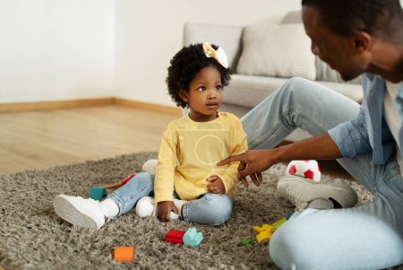 Photo for Loving african american dad teaching his toddler daughter to talk. Black man father sitting on carpet floor at home, communicating with his child little girl, playing with toys, copy space - Royalty Free Image