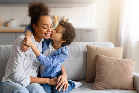 Téléchargez les photos : Preteen garçon noir en chemise bleue embrassant affectueusement sa joue de mères heureuses, mettant en valeur leur relation d'amour dans un cadre confortable à la maison, espace libre - en image libre de droit