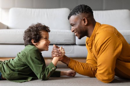 Foto de Entusiasta chico negro compite en un concurso de lucha libre de brazos con su padre, ambos mostrando grandes sonrisas, lo que indica diversión y vinculación de la actividad familiar en el interior, vista lateral - Imagen libre de derechos