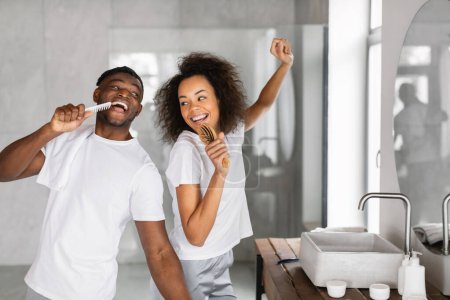 Téléchargez les photos : Jeune couple noir appréciant brosser les dents ensemble et s'amuser tout en chantant tenant brosses à dents comme des microphones, matin en famille dans l'intérieur de la salle de bain moderne. Soins dentaires - en image libre de droit