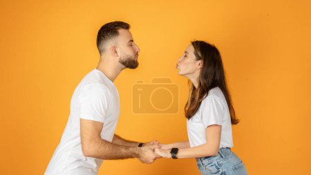 Téléchargez les photos : Un jeune couple européen joyeux et ludique tenant la main et se levant comme pour se préparer à un baiser, les deux vêtus de t-shirts blancs sur un fond orange vif, studio, panorama - en image libre de droit