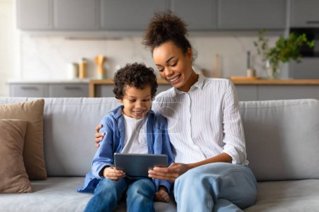 Foto de Alegre madre negra y su hijo preadolescente comparten un momento divertido mientras usan la tableta digital, sentados en un cómodo sofá en una sala de estar moderna y bien iluminada - Imagen libre de derechos