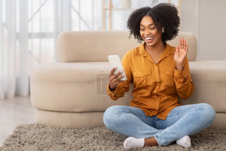 Téléchargez les photos : Joyeuse femme afro-américaine millénaire portant une chemise en moutarde et un jean bleu assis les jambes croisées sur un tapis, agitant la main tandis que la vidéo appelant sur son smartphone, profiter d'un appel vidéo - en image libre de droit